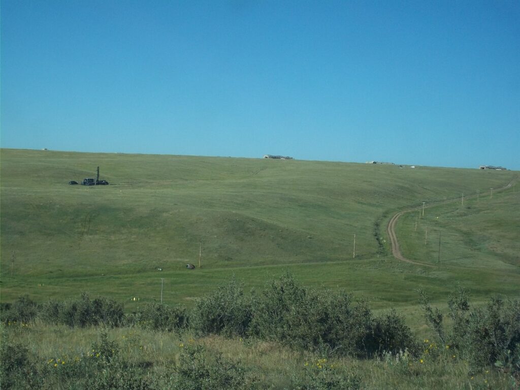 Photograph 7. This photo shows the western Transition Area looking southwest towards the Atlas Site 4 LSB Area – the missile site buildings are visible at the top of the slope. A Sonic drilling rig is visible in the middle distance at left, at the MW70 RI nested monitoring well location, installed in 2016. Lone Tree Creek flows from right to left (eastward) at the base of the slope in a small incised channel. The northeast groundwater TCE plume vector extending from LSB 2 flows towards the viewer below the unpaved road at right and the slope in the middle of the photo.